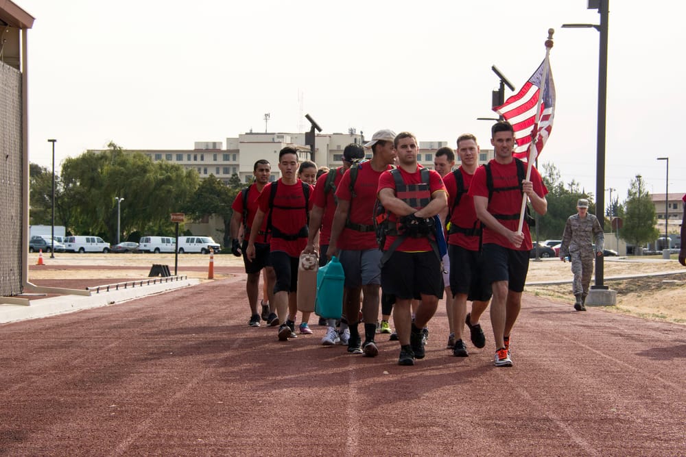 A group of GORUCK Light participants in matching red shirts and rucksacks walking together on a running track, one carrying an American flag.
