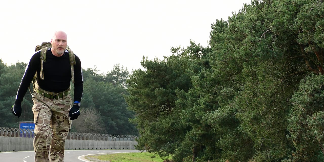 A man rucking alone along a paved road with a loaded pack on his back, trees lining the roadside.