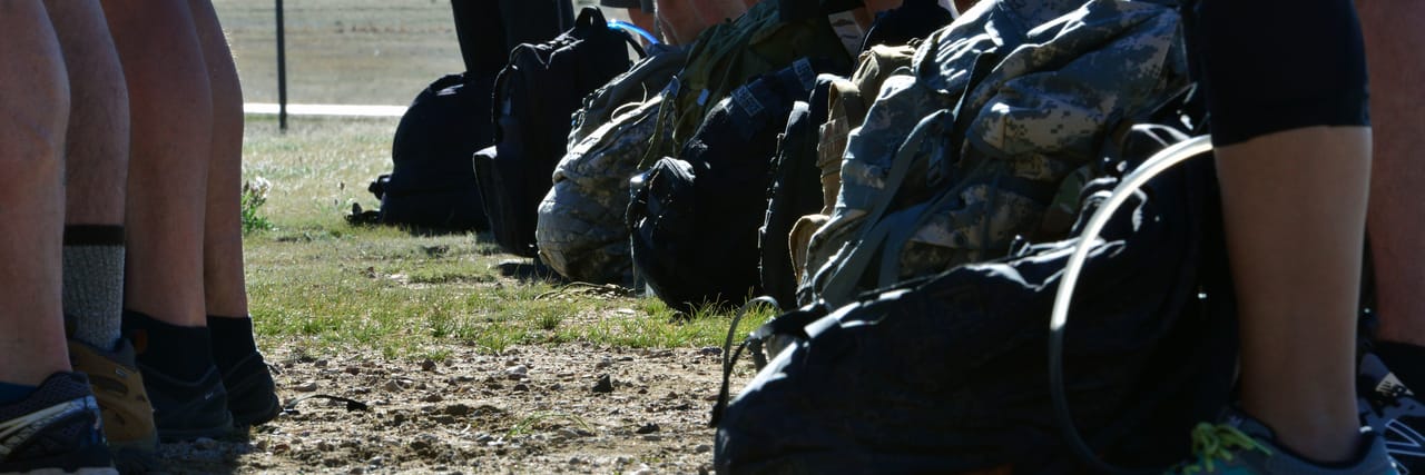 A row of loaded rucksacks resting on the ground at the feet of participants lined up before a ruck event.