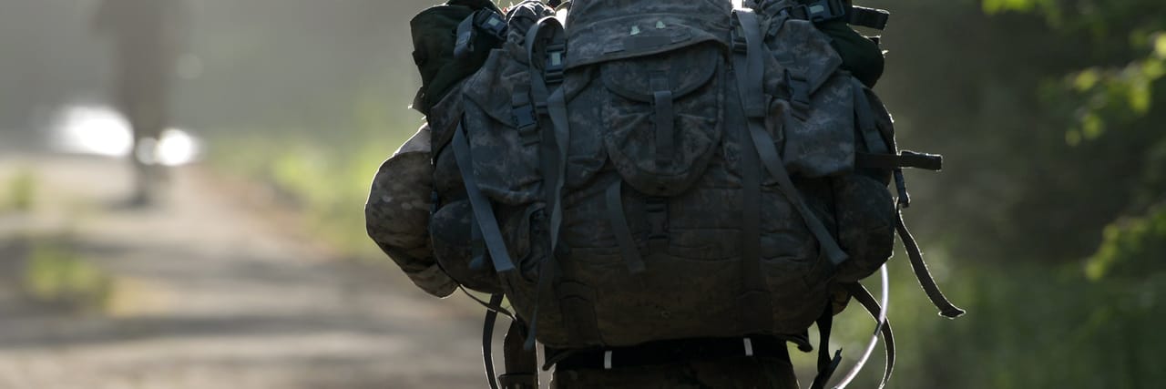 Close view of a heavily loaded rucksack on the back of a soldier walking a wooded trail in soft light.